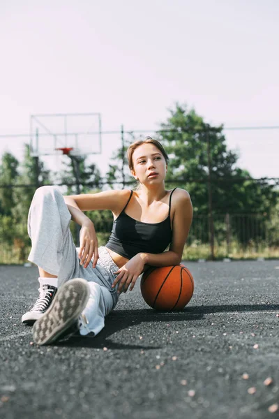 Potret seorang gadis cantik yang duduk di lapangan olahraga di taman atau sekolah dengan bola basket setelah pertandingan atau olahraga — Stok Foto