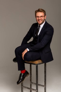 Siyah takım elbiseli, beyaz gömlekli, kravatlı, siyah çizmeli ve gözlüklü bir adam yüksek bir sandalyede oturuyor ve gülümsüyor. İş kıyafetleri. Bir adamın portresi. Başarı.