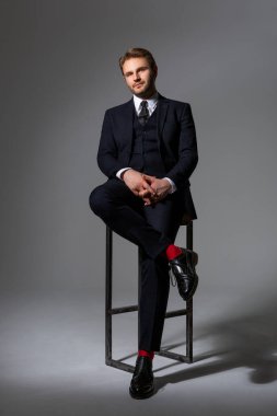 Siyah takım elbiseli, beyaz gömlekli, kravatlı, siyah çizmeli bir adam yüksek bir sandalyede oturuyor. Kendine güvenen bir bakış. İş kıyafetleri. Bir adamın portresi. Başarı.