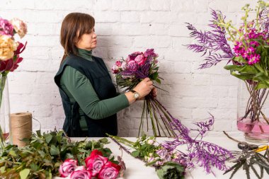 Beyaz tuğladan bir duvarın arkasında çiçekçi bir işyeri. Çiçekçi bir buket gül, karanfil ve okaliptüs yapar, adım adım çiçek ekler. Öğretici.