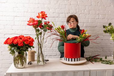 Beyaz tuğladan bir duvarın arkasında çiçekçi bir işyeri. Deneyimli bir çiçekçi aranjmanın tabanına kırmızı bir kutuda çiçek yerleştirir. Adım adım. Öğretici.