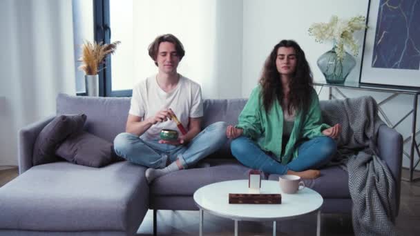 Pareja joven aprendiendo a cuidar la salud mental. Familia intenta meditar con un cuenco tibetano, de pie campana. Meditación con aplicación móvil. cámara lenta 4k — Vídeos de Stock