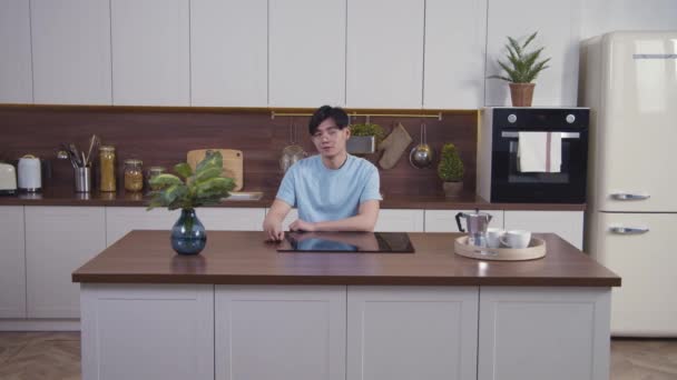 Portrait of Asian calm man smiling and sitting at modern kitchen, looking at camera. Medium shot, slow motion 4k — Stock Video