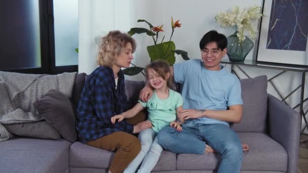 Багаторасовий сімейний портрет на дивані. Тато і дочка з фетро-фігурними малюнками на руках. Молодий азіатський батько і кавказька мати з дитиною розважається у вітальні — стокове відео