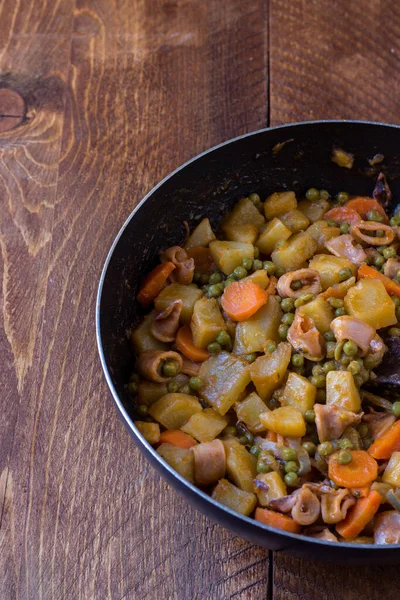 Ahşap arka planda tavada pişmiş patates, bezelye ve havuçlu kalamar.