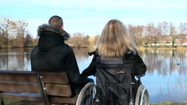 障害者の概念 隣のベンチに座っている男に手を差し出す車椅子の金髪の女性 湖に面した風景を考えるカップル — ストック動画