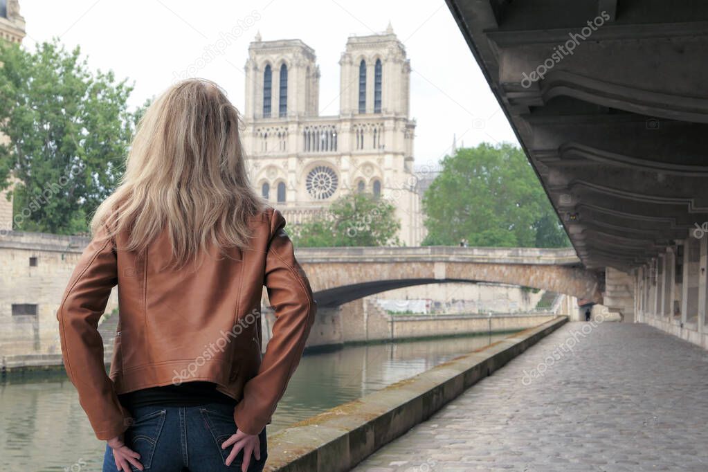 Blonde woman in trousers, standing from behind on the banks of the banks of the Seine. Cathedral of Notre Dame de Paris deliberately blurred in the background.