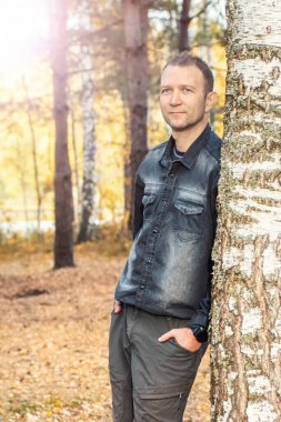 Bir adam sonbahar ormanında bir ağaca yaslanıyor, güneşin ışınlarındaki sonbahar manzarasına bakıyor..