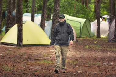 Nehir kıyısındaki ormanda kamp çadırlarının yanında yürüyen sert adam. Açık hava kamp konsepti