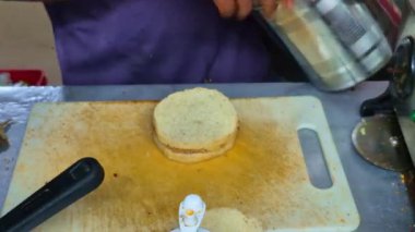 Bir adam peynirli burger hazırlarken görülüyor. İçindekiler düzgünce sıralanmış, gelişigüzel bir yemek pişirme anı sunuyor..