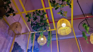 Yukarı bakan bir görüntü sarı geometrik tavan çerçeveleri içinde parlayan sıcak kolye ışıklarını ortaya çıkarır, sarkık yeşil bitkiler ve canlı, çağdaş bir iç atmosfer yaratan yapısal elementlerle çevrili..