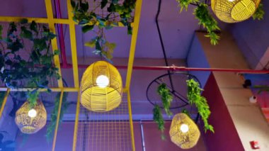 Ilık örülmüş kolye ışıkları sarı tavan çerçevelerini aydınlatıyor. Asılı yeşil yapraklar ve açık kırmızı borular ile vurgulanıyor. Katmanlı dokular ve açık renkli ve modern bir iç mekan ortamı yaratıyor..