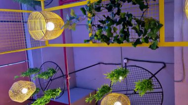 Asılı yeşil bitkiler, metal ağ aksanları ve görünür kablolar ile süslenmiş sarı ızgara tavanda ışıldayan sıcak kolye ışıkları, katmanlı derinlik ile canlı bir modern iç yaratıyor..