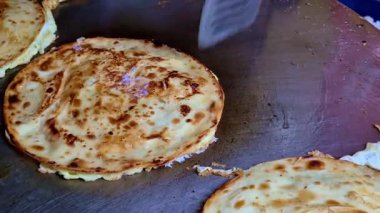 Büyük, düz bir ızgarada pişen çoklu eggroll parathas yakın plan, eşit kahverengili yüzeyler, pullu katmanlar ve çevirmeye hazır bir metal spatula.