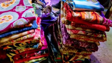 Üst düzey katlanmış, desenli battaniyelerden oluşan canlı bir pazar sahnesi. Renkli yün kumaşlar parlak ışık altında çeşitli çiçek ve geometrik tasarımlar sergiliyor..