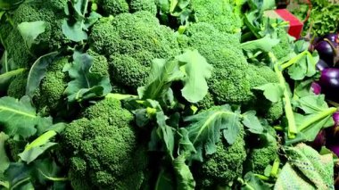 Taze brokoli çiçeklerinden oluşan büyük bir yığın çerçeveyi canlı yeşil renkleri ve engebeli dokularıyla doldurur. Çıtır yeşil yapraklar kafaların arasına saçılmış ve birkaç mor patlıcan yakınlarda oturuyor..