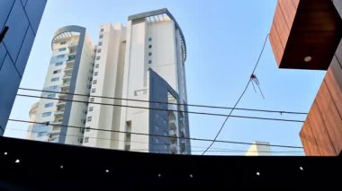 Parlak bir gökyüzüne karşı yükselen beyaz ve gri bir meskun bina. Birkaç siyah elektrik hattı ön tarafı çaprazlayarak üst katları kapatıyor..