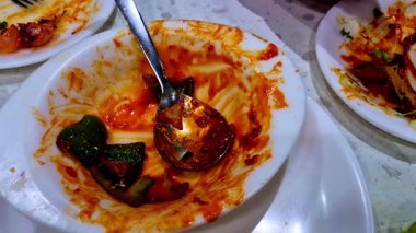 Yemekten sonra arta kalan kırmızı sos ve yeşil biber içeren beyaz bir kase. Bir metal kaşık dağınık kasenin içinde duruyor, baharatlı bir tabağın veya aperatifin kalıntılarını vurguluyor. Sahnede boş tabak var.