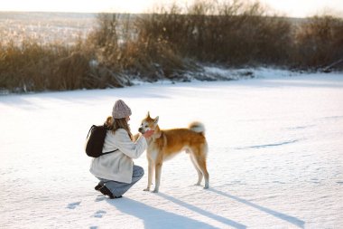 Karlı bir kış gününde Akita Inu köpeğine sarılan genç bir kadın. İnsan ve evcil hayvan arasındaki dostluğu, güveni ve sevgiyi gösteren sıcak duygusal bir an. Kopya alanı, doğal ışık, kış manzaralı yatay görüntü.