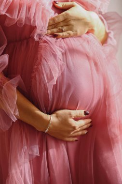 Yumuşak pembe tül elbise giymiş hamile bir kadının göbeğini nazikçe tutarken. Annelik, şefkat ve bebek konsepti.