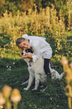 Küçük kız, altın saat boyunca köpeğine sarılıp, çayırda gülümsüyor. Sıcak duygusal bağ ve çocukluk arkadaşlığı kavramı