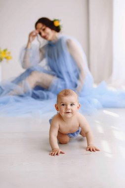 Yumuşak mavi elbiseli anne arka planda bulanık otururken bebek emeklerken derinlik ve hikaye anlatımı oluşturuyor. Annelik desteği, büyüme ve erken bağımsızlık kavramı.