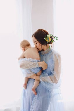 Çocuğu parlak bir pencerenin yanında kollarında tutarken, bebeğini nazikçe öpen genç bir annenin şefkatli portresi. Açık mavi şeffaf tül elbise ve çiçekli saç aksesuarı giyen bir kadın. Yumuşak doğal ışık havadar, duygusal bir anne...