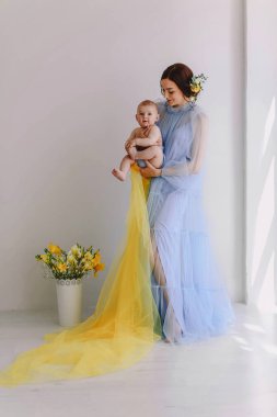 Yumuşak bir pastel mavi tül elbise giymiş gülümseyen genç anne sıcak bir etkileşim sırasında bebeğini nazikçe tutuyor. Parlak doğal ışık ve temiz iç mekan duygusal bağ, annelik bakımı ve modern ebeveynlik yaşam tarzı temaları vurgulanıyor.