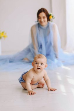 Arka planda zarif mavi bir tül elbise giymiş, gülümseyen bir anne izlerken, aydınlık bir zeminde sürünen meraklı bir bebek. Doğal gün ışığı ve minimal iç mekan otantik ebeveynlik, çocuk bakımı ve erken çocukluk gelişimi yaratır.