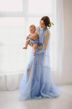 Yavrusunu büyük parlak bir pencerenin yanında tutan açık mavi tül bir elbise giyen genç bir anne. Yumuşak arka plan ve havadar iç mekan temiz bir duygusal annelik imajı yaratıyor ebeveynlik, aile ve çocuk bakımı reklamları için mükemmel..