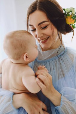 Bebeğini tutan hassas mavi tül bir elbise giyen gülümseyen genç bir annenin yakın duygusal portresi. Sıcak ifadeler ve hafif ışıklandırma güçlü bir annelik, ebeveynlik ve aile bağları kavramı yaratıyor. Reklamcılık için ideal.
