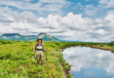 A fisherwoman dressed in waders holding a spinning rod and a fly rod ready to fish in a remote river in the  mountains of Alaska. clipart