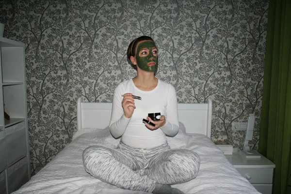 The girl sits on a bed with a green mask on her face. Home spa care.