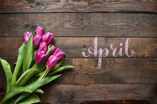 April lettering. Pink, tulips bunch on dark barn wood planks background. Postcard template.