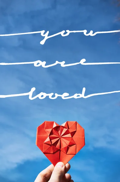 Corazón de papel de origami doblado rojo en la mano sobre el fondo azul del cielo. Plantilla de postal Hippster. Eres amado texto, letras . —  Fotos de Stock