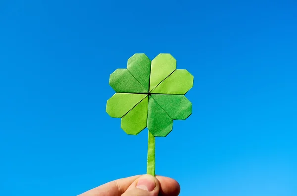 Hand holding green paper origami folded shamrock on blue sky background. Sunny weather outdoors. — Stock Fotó