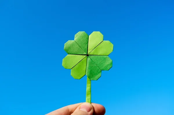 Mão segurando origami papel verde dobrado trevo no fundo do céu azul. Tempo ensolarado ao ar livre . — Fotografia de Stock