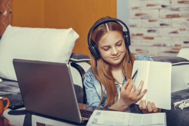 Kulaklıklı mutlu genç kız dizüstü bilgisayara bakarak notlar alıyor, kız öğrenci video konuşmalarıyla konuşuyor, öğretmenlik hocası web kamerası eğitimi alıyor, online eğitim, e-koçluk konsepti. 