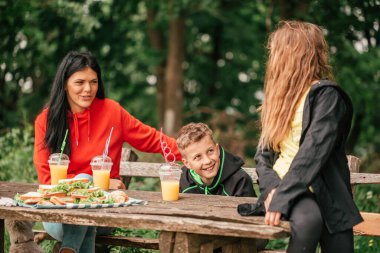 İki çocuklu güzel bir anne ormanda bir piknik masasında meyve suyu içip sandviç yiyor. Mutlu aile sohbetleri, doğanın tadını çıkarın ve birlikte eğlenin..