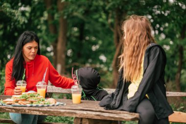 İki çocuklu güzel bir anne ormanda bir piknik masasında meyve suyu içip sandviç yiyor. Mutlu aile sohbetleri, doğanın tadını çıkarmak ve birlikte eğlenmek. Seçici odak.