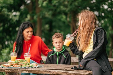 İki çocuklu güzel bir anne ormanda bir piknik masasında meyve suyu içip sandviç yiyor. Mutlu aile sohbetleri, doğanın tadını çıkarmak ve birlikte eğlenmek. Seçici odak.