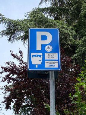 Açık gökyüzüne karşı yoğun yeşil ve kestane rengi yaprakların arasında İtalyan metinleriyle ödemeli park yerini gösteren açık mavi bir park yeri tabelası..