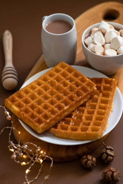 Winter sweet breakfast with waffles, honey, cocoa drink with marshmallows, served on a wooden board and a brown background with festive Christmas decorations. Winter mood. Close-up. Selective focus