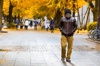 Kahverengi ceketli Asyalı bir adam elini kolunu sallaya bir parkta sonbaharda dönüyor