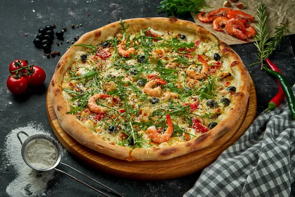 Oven-baked Italian pizza with sauce, cheese, arugula, shrimps and olives in a composition with ingredients on a dark background. Top view.Pizza with seafood