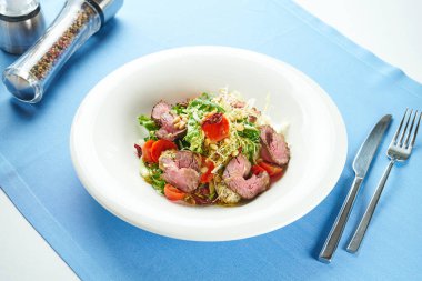 İştah açıcı, kızarmış biftekli sıcak salata, vişneli domates, parmesan ve çam fıstığı mavi masa örtüsünde beyaz bir tabakta.