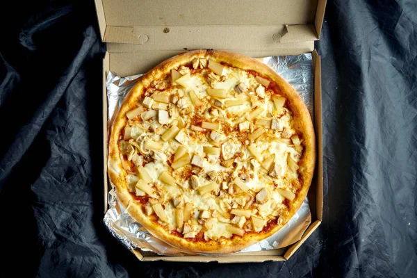 Crispy italian pizza with pineapple, melted cheese and chicken in a delivery package on a black background. View from above. Hawaiian pizza from the oven