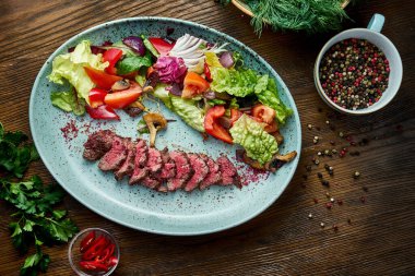 Orta pişmiş sığır etli ve taze sebzeli sıcak salata, ahşap arka planda mavi bir tabakta servis ediliyor. Restoran yemeği. Yaklaş, seçici odaklanma