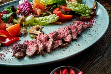 Orta pişmiş sığır etli ve taze sebzeli sıcak salata, ahşap arka planda mavi bir tabakta servis ediliyor. Restoran yemeği. Yaklaş, seçici odaklanma