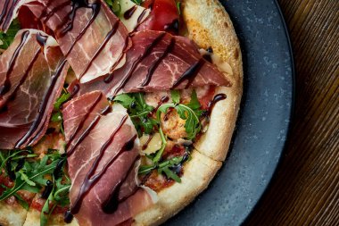 Jamon, roka, kırmızı sos ve gevrek kenarlı balzamik kaplı ahşap bir arka planda siyah bir tabakta servis edilen ahşap kaplı pizza. Bir çeşit İtalyan pizzası.